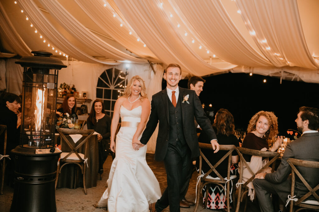 Photo of a bride and groom in a tent reception holding hands and celebrating their wedding.