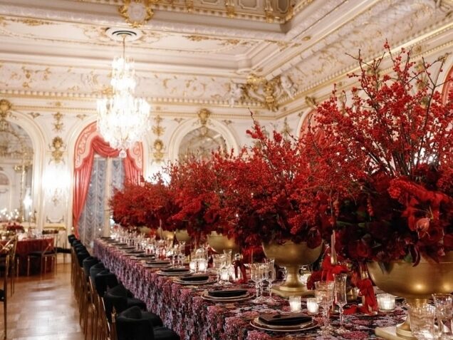 Photo By Lauryn Prattes Washington DC wedding planners of her reception area with large red flowers in gold vases on the long table that is adorned with floral linen and glassware and plates in a huge room with gold carvings.