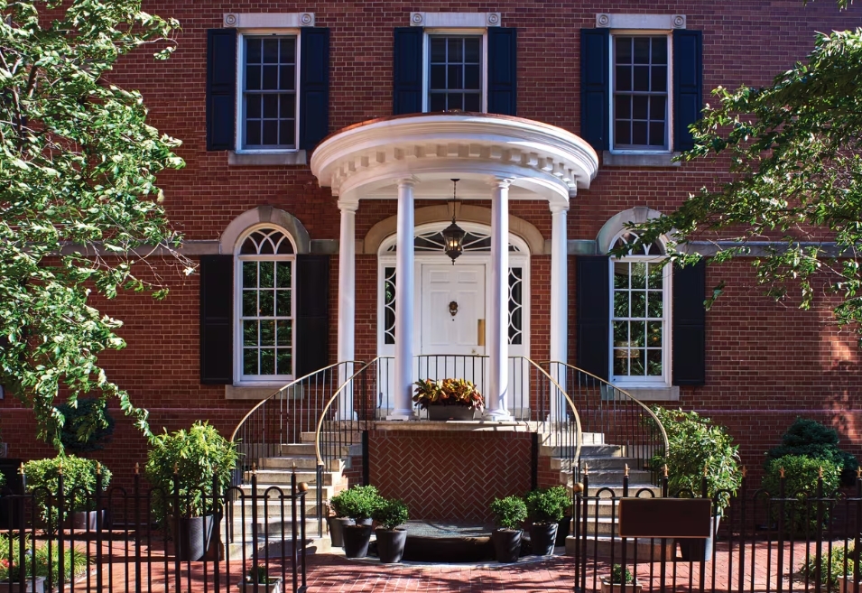 Photo the facade of the Morrison House with red brick and large columns at the front door with spiral stairs leading to entrance. Lush greenery and trees. 