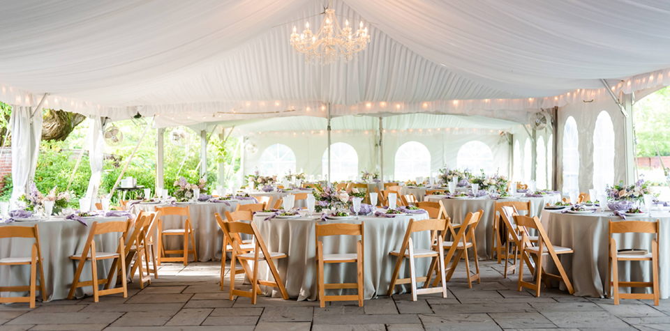 Photo of the reception area of The River Farm under a tent with tables adorned with table linen, floral, glass stemware and china. The tent is lined with a sheer drapery at the ceiling with a chandelier. Alexandria Virginia wedding Venue.