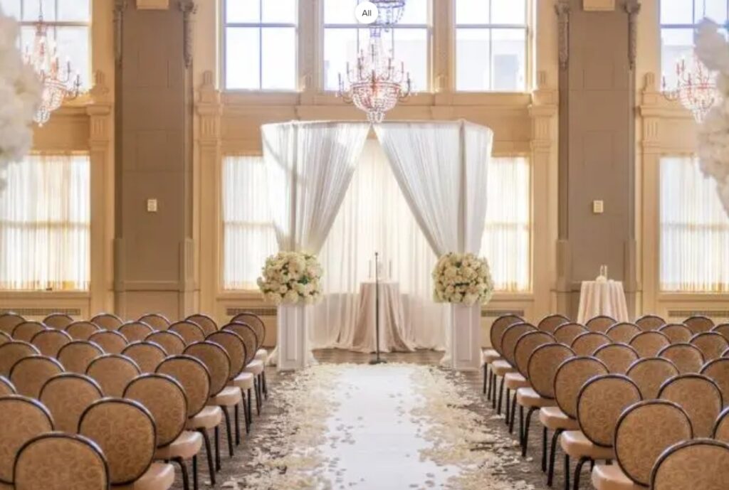 Photo of the ceremony area in John Marshall Ballrooms with rows of chairs and alter in white sheer material flanking the sheers are large florals. Large window let sunshine into the ceremony. A Richmond Virginia wedding venue. 