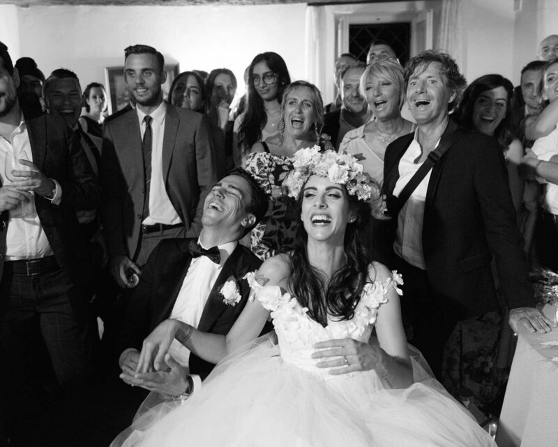 Photo by Pat Cori of a couple lauging and celebrating their vows. Bride and groom dressed in wedding attire sitting while family are circled around them. 