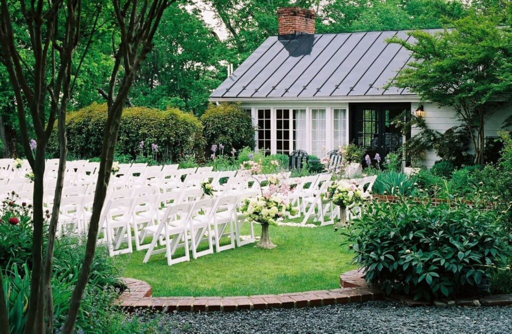 Photo of a outside ceremony setup at the Clifton Venue. Rows of white chairs with florals flanking the pathway. Surrounded by lush green lawn and trees. The back area of the Venue. This wedding venues Charlotte VA.