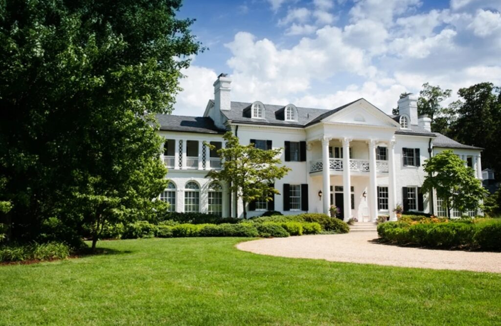 Photo of Keswick vineyard estate house with large columns flanking the entrance with black shutters on the windows surrounded by lush greenery. A wedding venue Charlottesville VA.