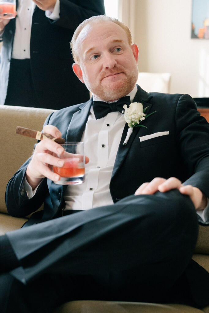Groom relaxing with a cigar and cocktail while getting ready for a black tie wedding at The Virginian Hotel in Lynchburg, Virginia.