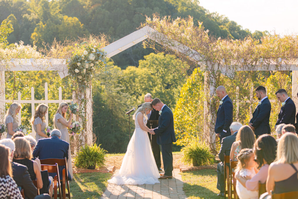 Outdoor wedding ceremony in Virginia beneath a white arbor covered in greenery, bride and groom exchanging vows surrounded by close family and friends, photographed by Pat Cori Photography for a blog on wedding venues for small weddings