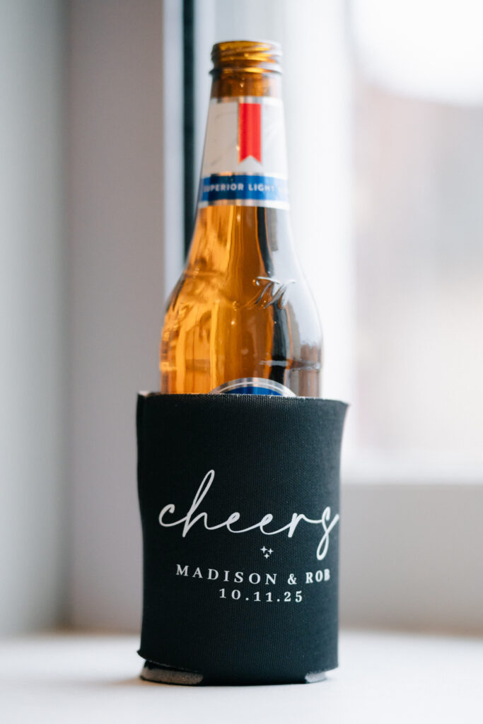 Personalized beer koozie reading ‘cheers Madison & Rob 10.11.25’ displayed during cocktail hour at The Virginian Hotel wedding