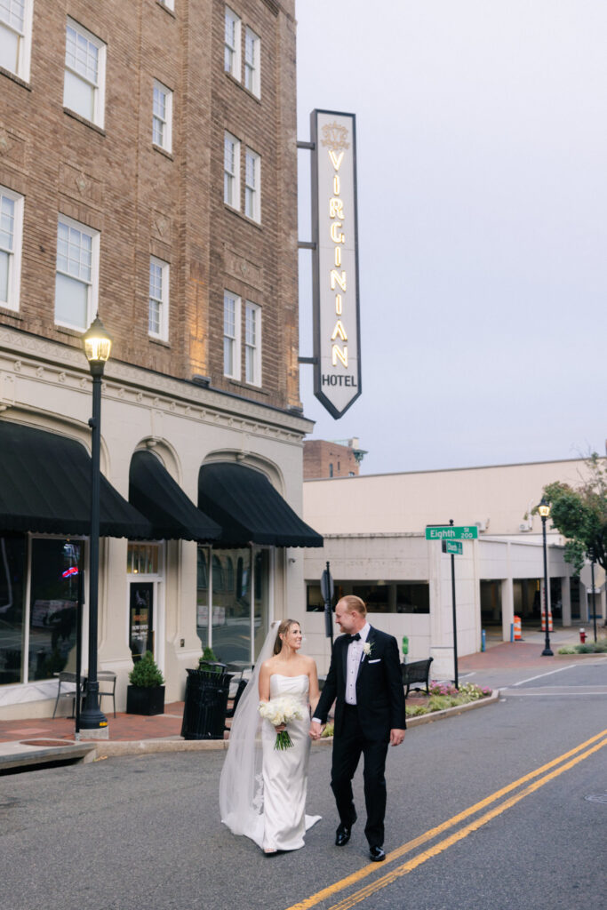 Bride and groom walking hand in hand down the street beside The Virginian Hotel sign in downtown Lynchburg during sunset portraits photographed by Pat Cori Photography