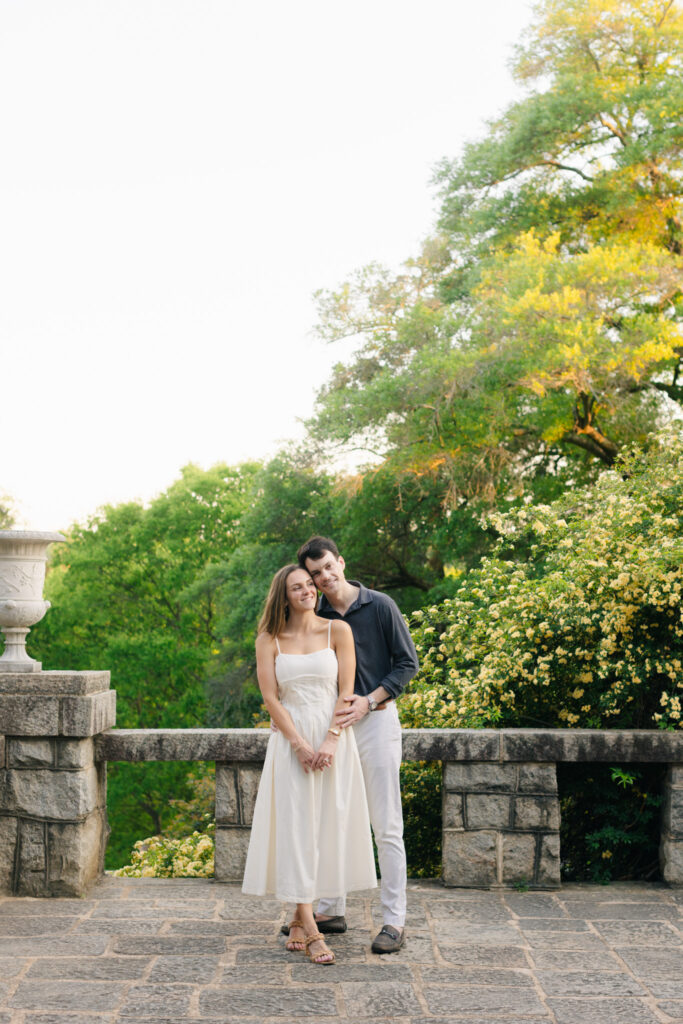 Elegant engaged couple standing together on a stone terrace at Maymont Park in Richmond Virginia, dressed in soft neutral tones with lush greenery behind them