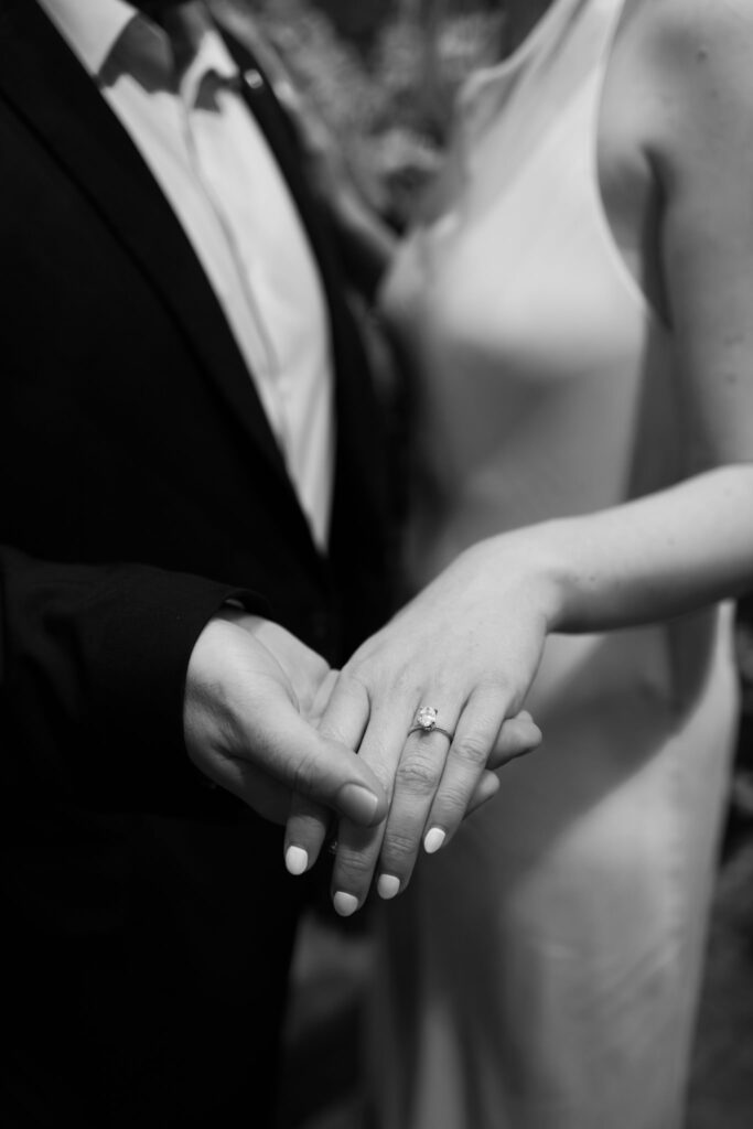 Black-and-white close-up of a bride and groom holding hands with her engagement ring in focus.