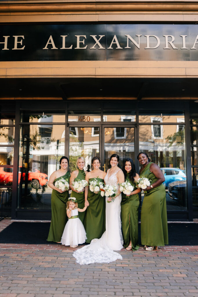 Bride and bridesmaids in olive green dresses holding white floral bouquets outside The Alexandrian in Old Town Alexandria.