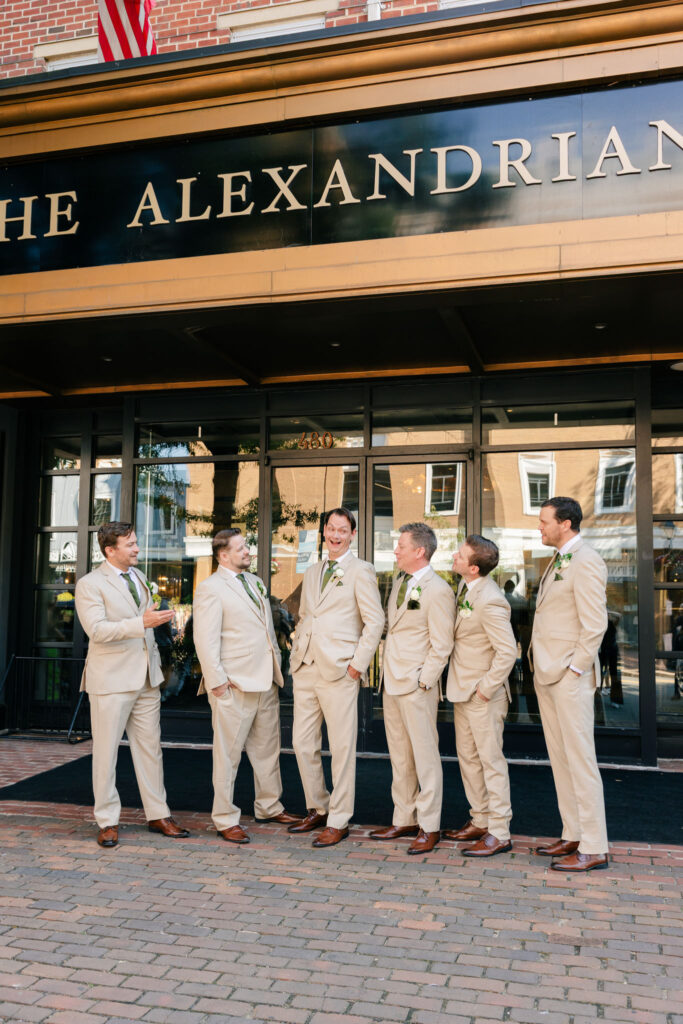 Groom and groomsmen in light neutral suits with green ties laughing together outside The Alexandrian Hotel.