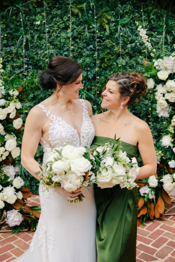Bride and bridesmaid holding white rose bouquets in The Alexandrian courtyard, surrounded by greenery and white floral arrangements