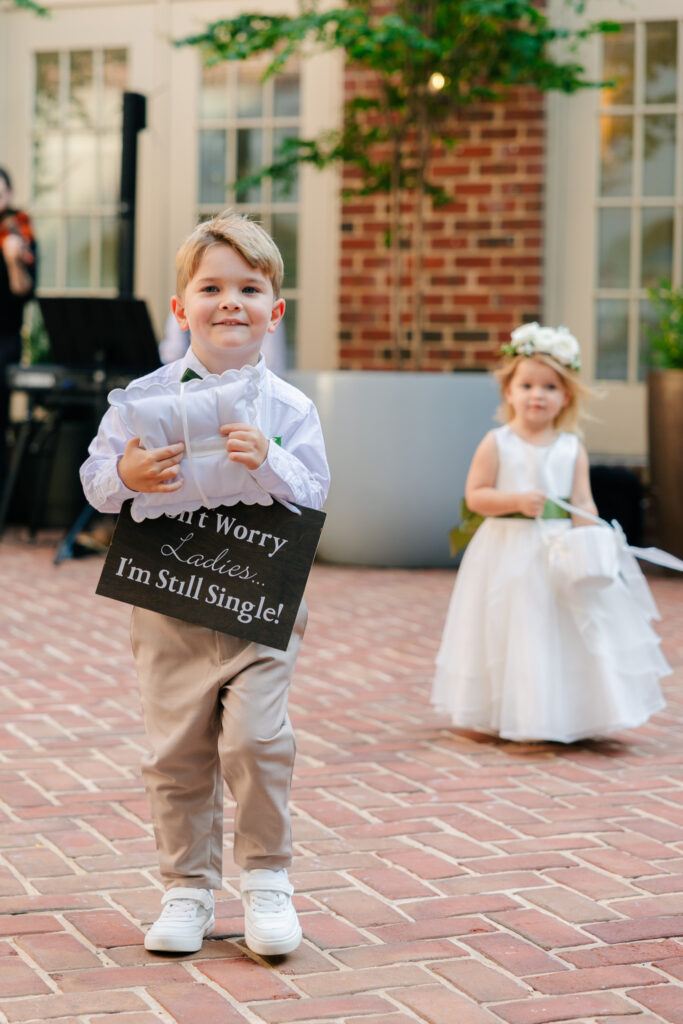 oung ring bearer carrying a ‘Don’t Worry Ladies, I’m Still Single’ sign with flower girl walking behind him during an outdoor wedding ceremony.
