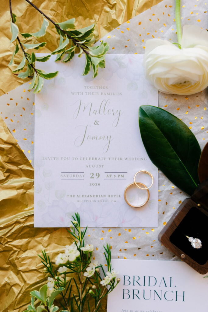 Alexandrian Hotel wedding invitation with gold wedding rings, white flowers and magnolia leaves in an elegant green, white and gold flat lay