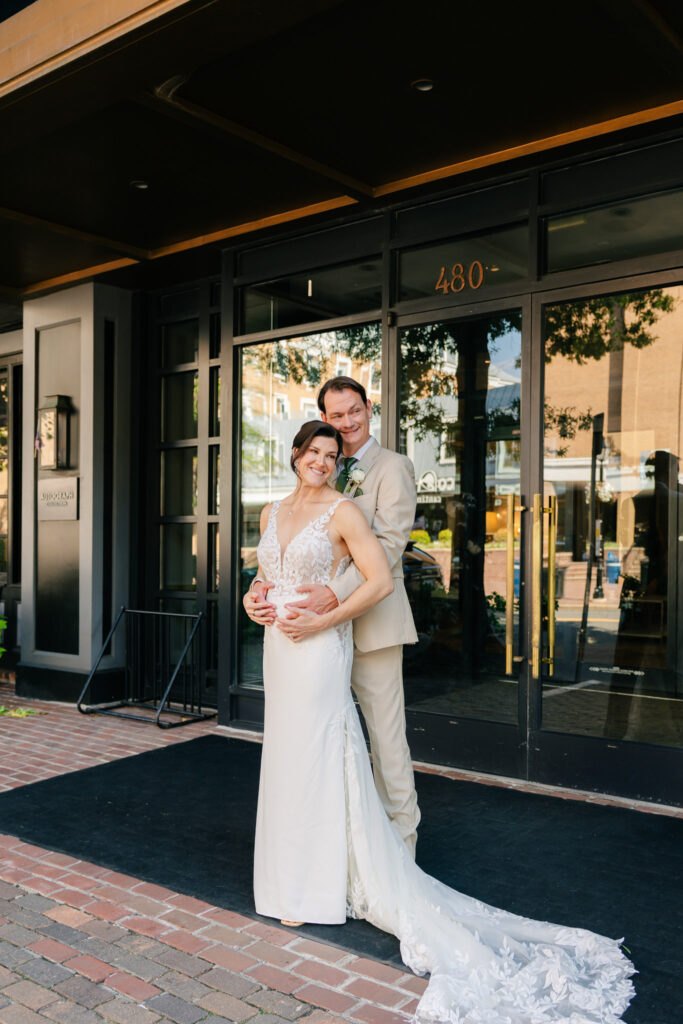 Bride and groom embracing outside The Alexandrian Hotel, with the bride’s lace wedding gown and long train on the brick sidewalk