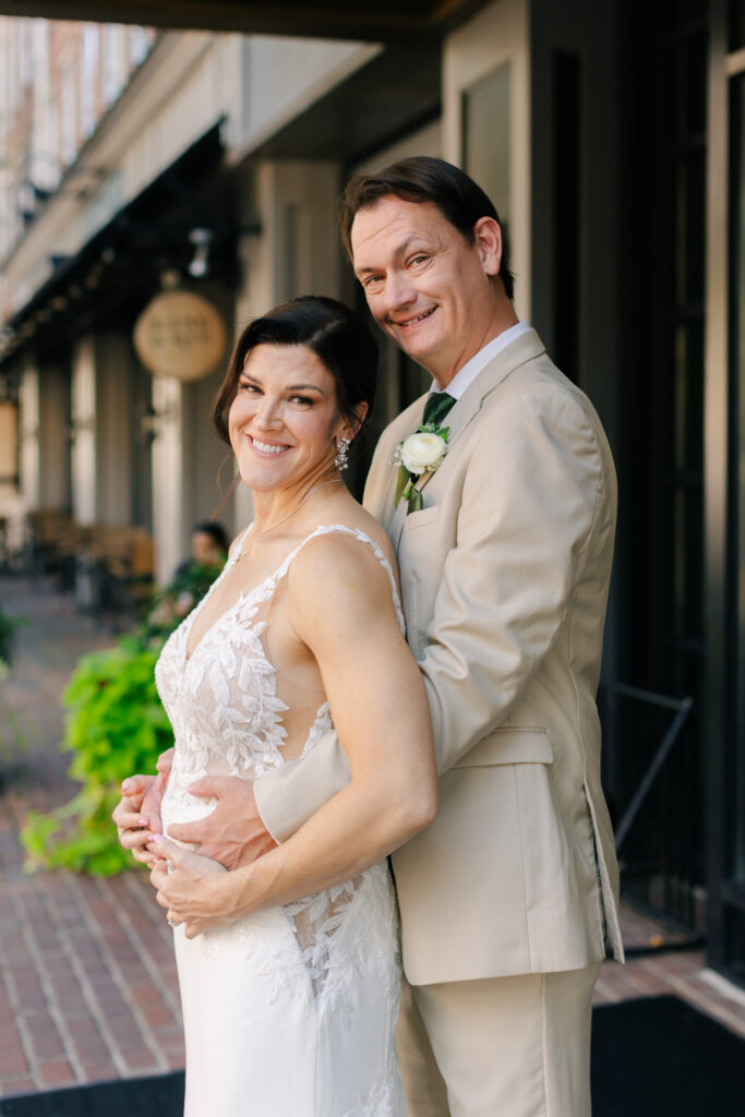 Smiling bride and groom posing together outside The Alexandrian in Old Town Alexandria, with the groom wearing a neutral suit and green tie