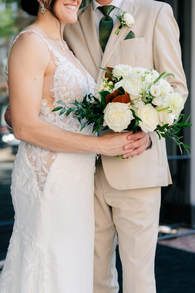 Close-up of a bride holding a white floral wedding bouquet with roses, greenery and magnolia leaves beside the groom in a neutral suit