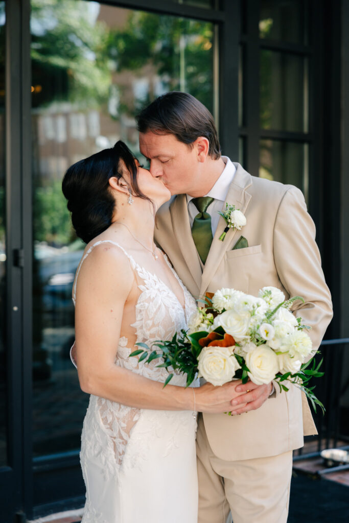 Bride and groom kissing outside The Alexandrian while holding a white rose and greenery wedding bouquet.